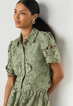 Žena nosí krátky rukáv olivovo zelenú čipkovanú blúzku s kvetinovými vzormi a zlatými gombíkmi, doplnenú zlatými náušnicami v tvare srdca.