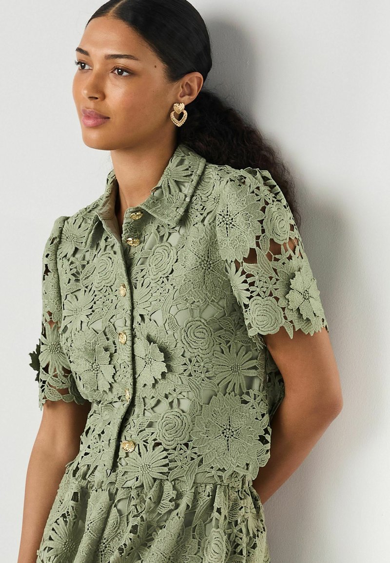 Femme portant un chemisier en dentelle vert olive à manches courtes avec des motifs floraux et des boutons dorés, associé à des boucles d'oreilles dorées en forme de cœur.