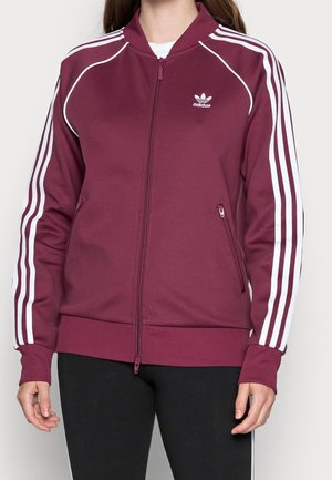 Person wearing a zipped maroon Adidas track jacket with white stripes on sleeves and logo on the chest, paired with black pants.