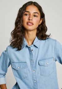 Lichtblauwe denimshirt met knoopsluiting aan de voorkant, twee borstzakken en opgerolde mouwen. Heeft witte knopen en subtiele stikseldetails.