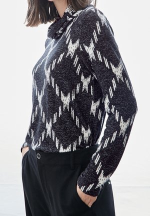 Femme portant un pull à manches longues noir et blanc à motif avec un col montant, mains dans les poches d’un pantalon noir, légèrement tournée vers la gauche.