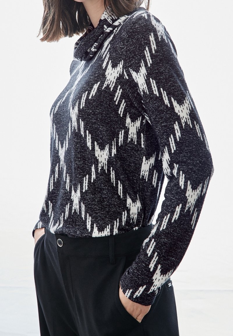 Femme portant un pull à manches longues noir et blanc à motif avec un col montant, mains dans les poches d’un pantalon noir, légèrement tournée vers la gauche.