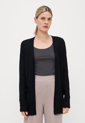 Zwarte lange mouwen cardigan gemaakt van zachte stof met een open voorkant, geribbelde manchetten en twee voorkzakken, gedragen over een grijze top en lichtroze broeken.