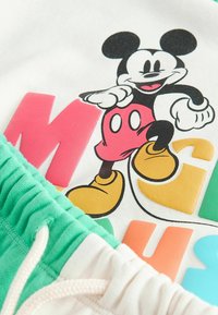 Micky Maus Cartoon mit roter Shorts und gelben Schuhen, gedruckt auf weißem Stoff mit buntem, teilweise sichtbar Text und grünem elastischem Bund darunter.