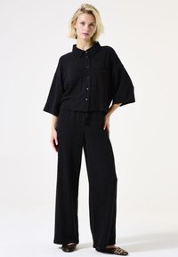 Schwarzes, strukturiertes Zweiteiler-Outfit bestehend aus einem kragenlosen Hemd mit Knöpfen und weiter Hose, beide mit lockerer Passform und minimaler Gestaltung.