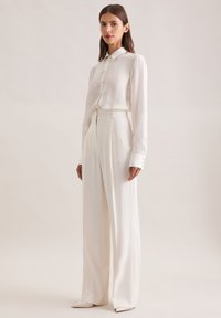 Witte blouse met lange mouwen en een knoopsluiting aan de voorkant, gecombineerd met hooggesneden, wijde witte broek en schoenen met een puntige neus. Gladde textuur.