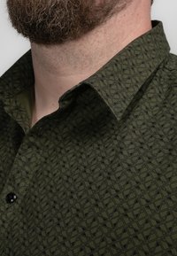 Camisa verde con un diseño geométrico, cuello puntiagudo y botones negros. La tela texturizada complementa el diseño general.