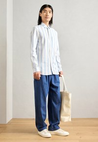 Chemise à boutons avec des rayures verticales bleu clair et blanches, assortie à un pantalon en denim bleu ample et des baskets blanches ; portant un sac fourre-tout beige.
