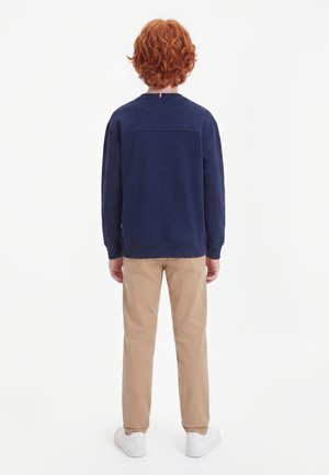 Enfant aux cheveux roux bouclés portant un sweat-shirt bleu marine, un pantalon beige et des baskets blanches, debout de dos sur un fond blanc.