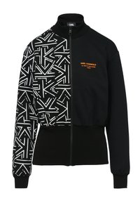 Veste zippée noire avec un design à moitié imprimé, présentant des formes géométriques blanches d'un côté et un noir uni de l'autre. Accents de logo orange.