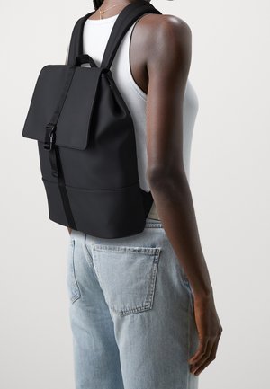Sac à dos noir avec rabat et boucle porté par une personne en débardeur blanc et jeans bleu clair, vu de côté et de dos.