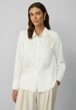 White button-up shirt with long sleeves, featuring gold embroidered accents resembling floral patterns on the left side. Smooth texture.