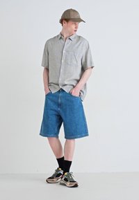 Chemise à manches courtes gris clair, shorts en denim bleu, chaussettes noires et baskets multicolores ; look décontracté avec une silhouette relaxée.
