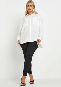 Camicia bianca con maniche lunghe e due tasche sul petto, abbinata a jeans skinny neri e sandali con tacco a stiletto.