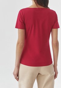 Nainen, jolla on päällään yksinkertainen punainen lyhythihainen paita ja beigehousut, näkyy selkäpuolelta valkoista taustaa vasten.