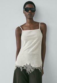 Frau mit kurzem Haar, die schwarze Sonnenbrille, ein weißes ärmelloses Oberteil mit Spitzenabschluss und schwarze Hose vor einem schlichten, hellen Hintergrund trägt.