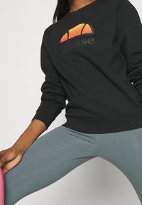 Personne portant un sweat-shirt noir avec un logo orange et des leggings bleu clair, tenant un kettlebell rose, en position de fitness.