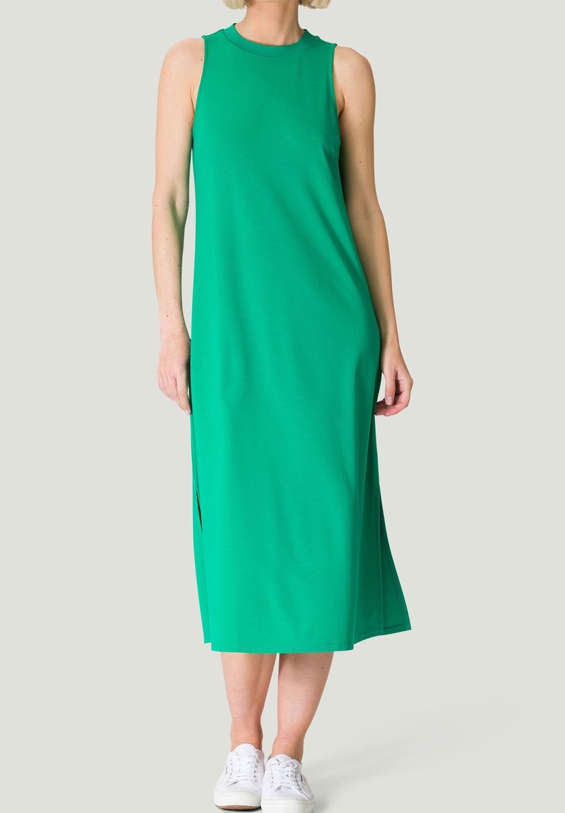 Femme portant une robe midi verte sans manches et des baskets blanches, debout devant un fond clair uni.