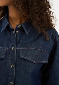 Traper košulja s ovratnikom na gumbe, tamnoplave boje, i dva džepa na prsima. Ima istaknuto zlatno šivanje i velike crne gumbe.