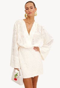 Vrouw met gladgestreken haar draagt een witte gebreide jurk met brede mouwen, gouden creolen, en houdt een wit handtasje vast met een kersenontwerp.