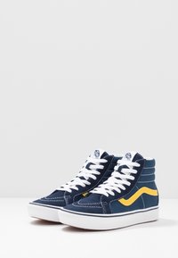 Vysoké tenisky vyrobené z tmavě modré plátna s bílými tkaničkami, žlutým bočním pruhem, bílou podrážkou a kontrastními prošívacími detaily.