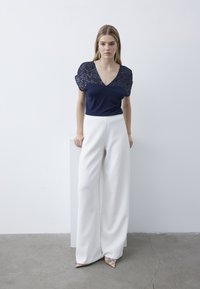 Top blu navy con maniche a cappuccio in paillettes e scollatura a V, abbinato a pantaloni bianchi a gamba larga e décolleté oro lucido a punta. Design minimalista, texture morbida.