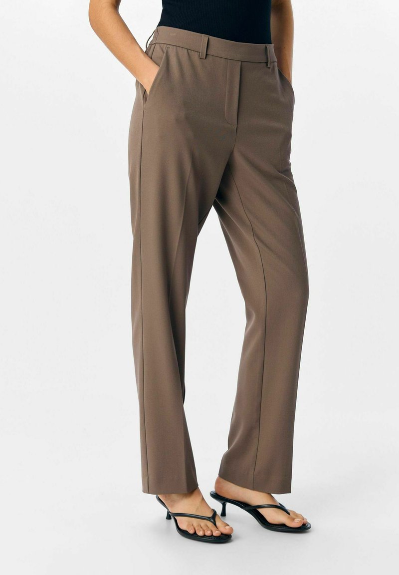 Personne portant un pantalon marron taille haute et jambes droites, les mains dans les poches, et des sandales à talons noirs sur fond blanc.