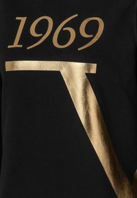 Svart t-shirt med metallic guldtryck som visar siffrorna "1969" och ett stiliserat bokstav "V" i en elegant design och mjuk textur.