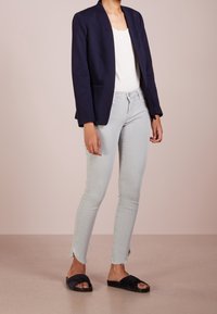 Personne portant un blazer bleu marine, un haut blanc, un jean gris clair raccourci avec des bords effilochés, et des sandales plates noires, debout sur un sol beige.