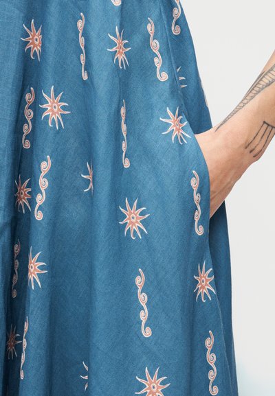 Χέρι με γεωμετρικά τατουάζ που ξεκουράζεται στην τσέπη μπλε υφάσματος με πορτοκαλί αστέρια και σχέδια στροβίλων.