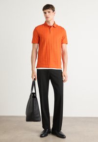 Camisa polo texturizada laranja de mangas curtas, combinada com calças pretas sob medida e sapatos de couro, carregando uma bolsa tote preta.