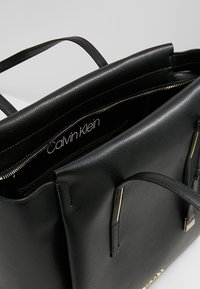 Musta Calvin Klein -nahkalaukku, jossa on kaksoiskahvat, avoin yläpää, joka paljastaa sisäisen vetoketjutaskun ja Calvin Klein -logon sisällä.