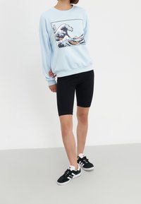 Sudadera azul claro con gráfico de olas, leggings negros y zapatillas negras. La sudadera tiene puños y dobladillo acanalados.