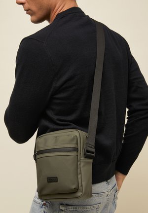 Mees, kes kannab musta kampsunit ja heledaid teksapükse, kandes oliivrohelist läbi keha kantavat kotti lukuga ees.