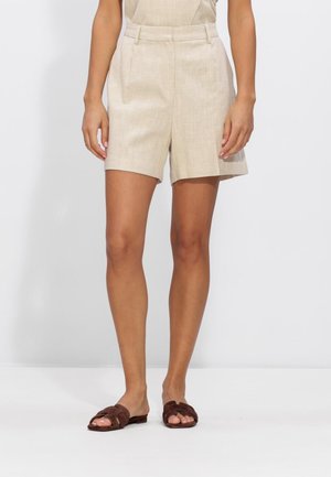 Vrouw die beige op maat gemaakte shorts met hoge taille en bruine platte slippers draagt, staand tegen een effen lichte achtergrond.