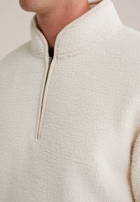 Pullover in pile crema con zip a metà davanti, colletto alto e tessuto morbido e testurizzato. Presenta un bordo beige lungo i lati della zip e del colletto.