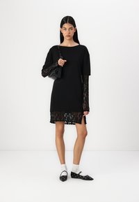 Fekete ruha rövid ujjal, csipke részletekkel az alján, fekete kézitáskával és cipőkkel, fehér bokazoknival, semleges háttér előtt.