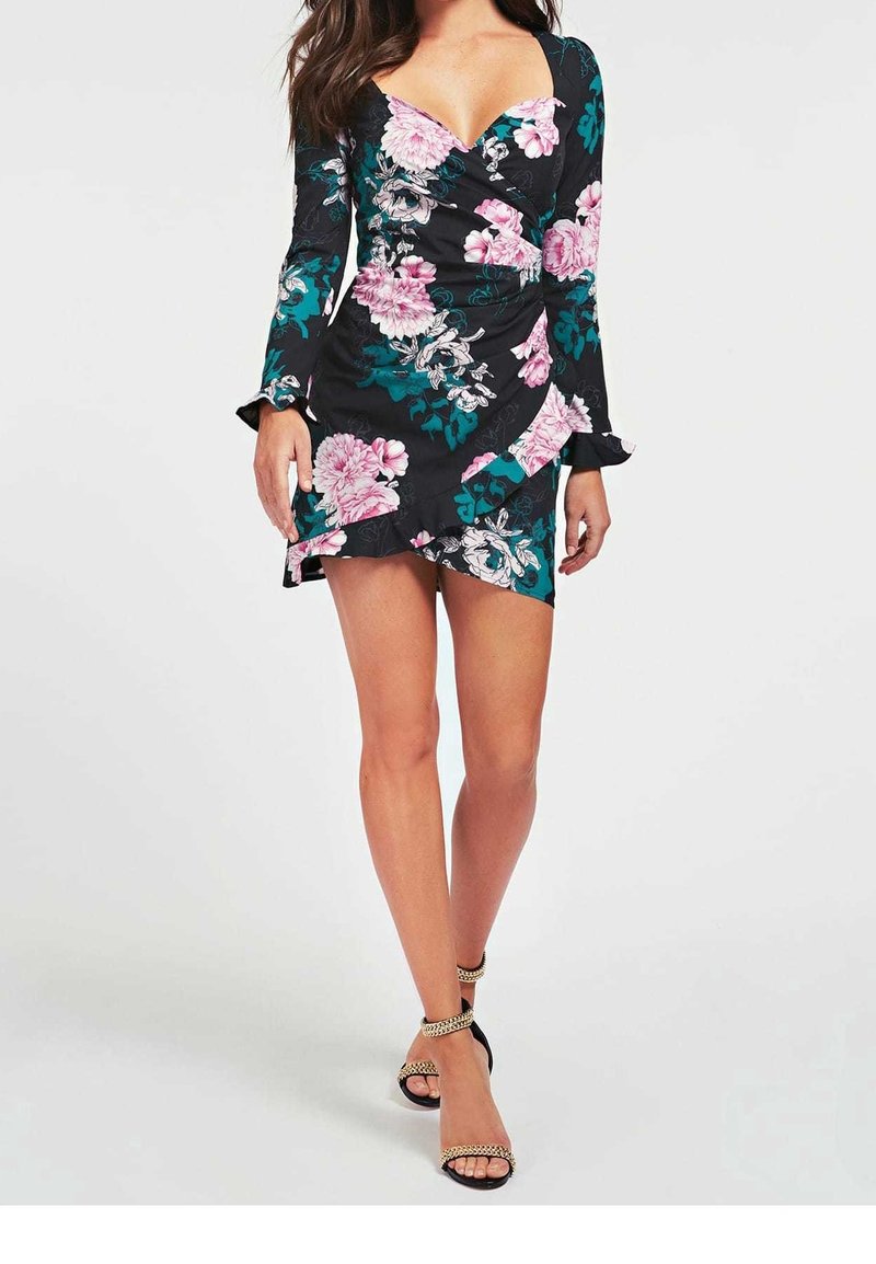 Femme portant une robe portefeuille noire à imprimé floral avec manches longues et ourlet volanté, assortie à des sandales à talons noirs avec détails en chaîne.