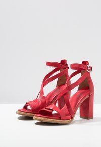 Rode leren sandalen met hoge hakken, enkelschoenen met bandjes en een blokhak, tentoongesteld op een witte ondergrond tegen een lichtgrijze achtergrond.