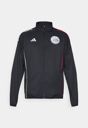 Veste de sport noire en matériau léger, avec des accents blancs et rouges, un col montant, une fermeture éclair complète et un logo rond sur la poitrine.