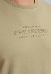 Kaki majica s okruglim izrezom, kratkim rukavima i utisnutim tekstom "FREEDOM UNLIMITED PME LEGEND AMERICAN CLASSIC" na prednjoj strani.