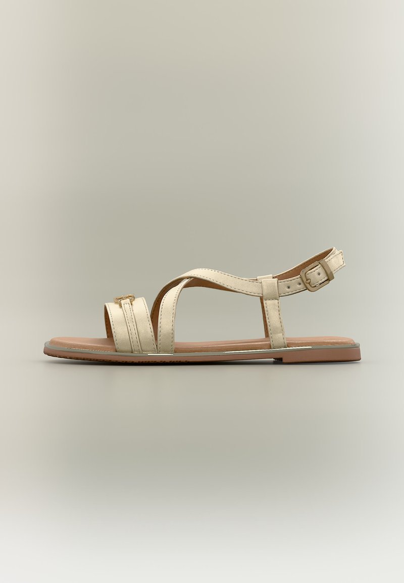 Μπεζ επίπεδη σάνδαλα με αγκράφα στον αστράγαλο, δύο μπροστινές λουρίδες και ανοιχτό δάκτυλο σε ανοιχτόχρωμο ουδέτερο φόντο.