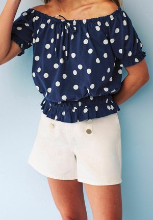 Femme portant un haut bleu marine à épaules dénudées avec des pois blancs et un short blanc à taille haute avec des boutons décoratifs.
