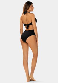 Kobieta stojąca na boso, zwrócona bokiem, nosząca czarny dwu-częściowy strój kąpielowy z przewiązanymi wstążkami z tyłu.