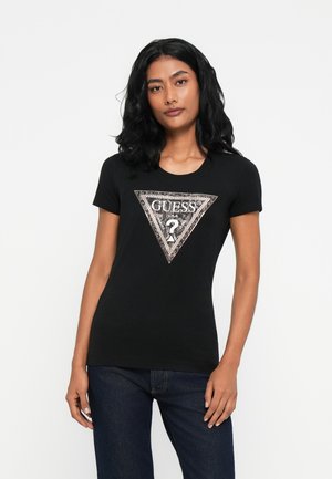 Donna in piedi con capelli neri lunghi e sciolti, che indossa una t-shirt nera di Guess con un logo a triangolo di paillettes e jeans blu scuro.