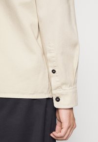 Poignet de chemise beige à manches longues avec deux boutons noirs au poignet au-dessus d'un pantalon noir, montrant une partie de la main et du bras d'une personne.