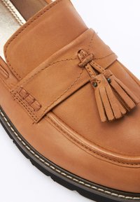 Mocassin en cuir tan avec un gland décoratif, présentant des accents brodés et une texture lisse. La semelle est noire avec un bord de couture contrasté.