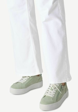 Gröna sneakers gjorda av texturerat material med vita snören, en dragkedjedetalj och en tjock vit sula, parat med vita raka byxor.
