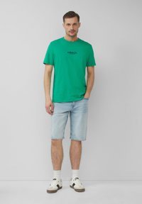 Groene katoenen T-shirt met een ronde halslijn en korte mouwen, voorzien van een zwart logo, gecombineerd met lichtdenim shorts en witte sneakers.
