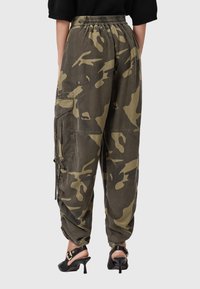 Persona che indossa pantaloni cargo larghi a vita alta con stampa camouflage verde e marrone e sandali neri con tacco e cinghie alla caviglia.